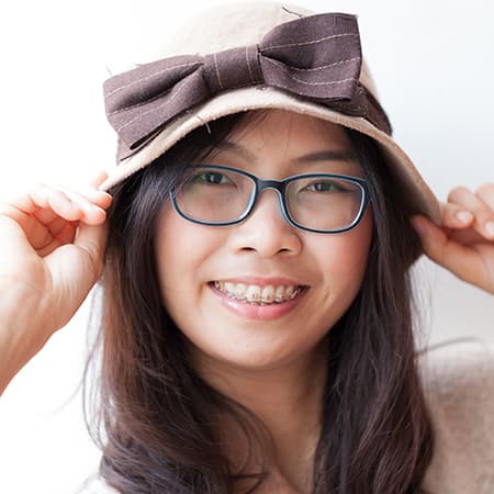 A Smiling lady with braces and eyeglasses and is wearing a hat with her hands holding the sides