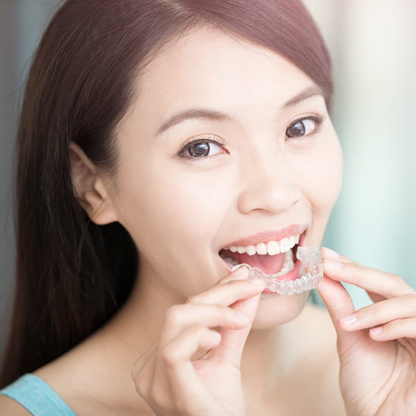 A smiling Asian lady with long hair is holding an invisalign close to her mouth